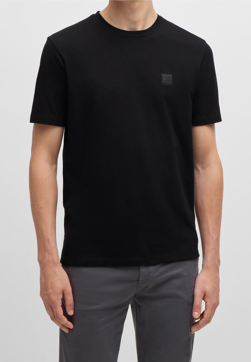 Homme portant un t-shirt noir uni à manches courtes avec un petit logo "BOSS" sur la poitrine et un pantalon gris sur un fond uni.
