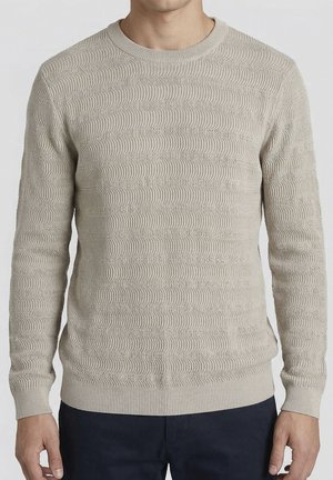 Maglione beige lavorato a maglia con collo rotondo indossato da una persona, con maniche lunghe, abbinato a pantaloni scuri.