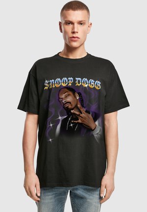 Nuori mies, jolla on musta t-paita, jossa on violetti kuvio ja teksti "SNOOP DOGG" Snoop Doggin muotokuvan yläpuolella, ja hän tekee käden elettä.