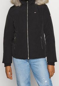 Veste noire isolée avec une capuche doublée de fourrure, fermeture éclair argentée et deux poches latérales ; présente des poignets côtelés et un logo sur la poitrine.