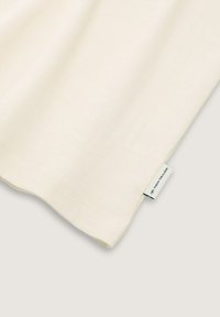 Camiseta de algodón color crema con textura acanalada y escote redondeado. Cuenta con una pequeña etiqueta de marca azul y blanca en el dobladillo.