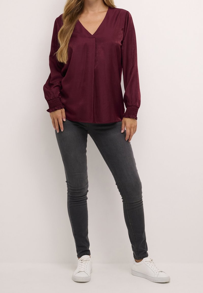 Femme portant un chemisier bordeaux à manches longues avec poignets froncés, un jean skinny gris foncé et des baskets blanches sur un fond uni.