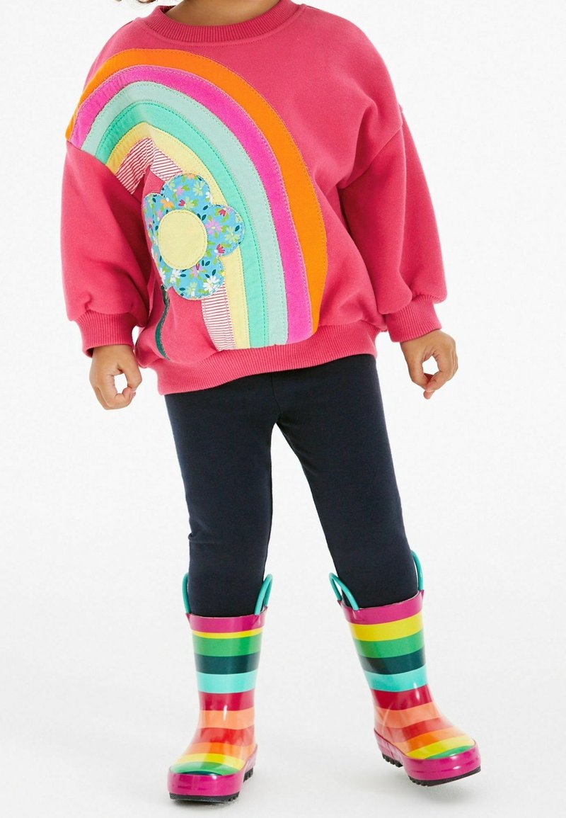 Enfant portant un pull rose avec un motif arc-en-ciel et fleur, des leggings noirs et des bottes de pluie rayées colorées, debout sur fond blanc.
