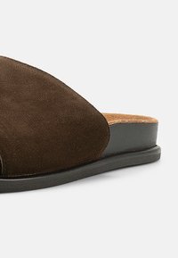 Καφέ σανδάλι από σουέντ με ευρύ πάνω λουρί, υφή στην επιφάνεια και αντίθετη μαύρη λαστιχένια σόλα. Απλός, μοντέρνος σχεδιασμός.