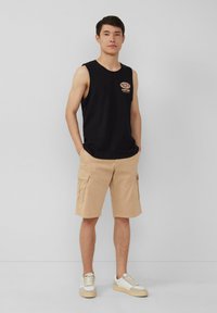 Schwarzes ärmelloses Tanktop mit "PACIFIC CUSTOMS" Druck; beige Cargo-Shorts mit Seitentaschen; weiße Sneaker mit grauen Akzenten.