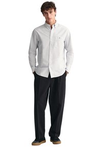 Camicia a maniche lunghe bianca con righe grigie, abbinata a pantaloni neri larghi e scarpe da ginnastica scure. La camicia presenta una tasca sul petto.