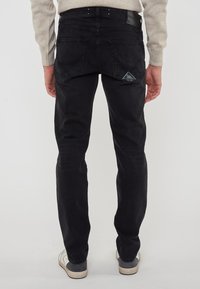 Persona che indossa jeans neri e sneakers bianche, in piedi con la schiena rivolta verso la fotocamera su uno sfondo semplice.