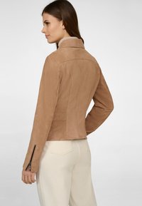 Brun mockaskinnjacka med en figursydd silhuett, hög krage och dragkedjor vid ärmarna. Den släta texturen framhäver den skräddarsydda designen.