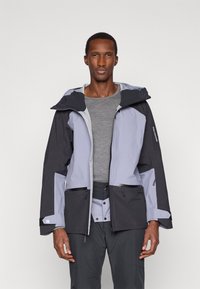 Homem alto vestido com uma jaqueta de exteriores cinza e preta de duas tonalidades sobre uma camisa cinza, posando contra um fundo claro liso.