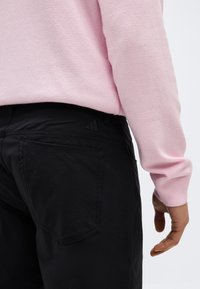 Άτομο που φοράει ανοιχτό ροζ μακρυμάνικη μπλούζα και μαύρο παντελόνι, εμφανιζόμενο από τη μέση και κάτω με κοντινή εικόνα της πίσω τσέπης.