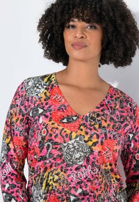 Une femme aux cheveux bouclés porte un haut V échancré vibrant orné de taches de léopard, de visages de tigre, d'yeux et de motifs colorés de style graffiti.
