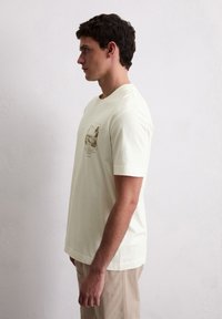 Tricou din bumbac de culoare crem, cu mâneci scurte, având un imprimeu grafic pe piept, asortat cu pantaloni bej deschis.