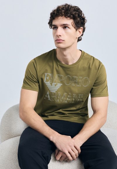 T-shirt vert olive avec logo "Emporio Armani" en gris clair. Manches courtes, col rond, matière en coton. Assis sur une chaise claire texturée.