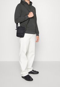 Bolso cruzado negro con dos bolsillos frontales, llevado sobre una sudadera oscura y pantalones blancos, acompañado de zapatillas negras en un fondo neutral.