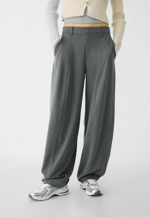 Person trägt weite graue Hosen, silberne Sneaker, ein kurzes graues Oberteil und eine cremefarbene, gerippte Strickjacke mit Händen in den Taschen.