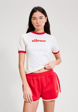 Ung kvinde iført hvid t-shirt med rød kant og "ellesse"-logo, kombineret med røde træningsshorts, stående mod en lys baggrund.