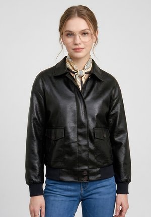 Jeune femme portant des lunettes transparentes, un foulard à motifs autour du cou, une veste en cuir noire et un jean bleu sur un fond uni.