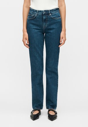 Nő, aki magas derekú, egyenes szárú kék farmert, fehér ujjatlan felsőt és fekete orrú Mary Jane cipőt visel, fehér háttér előtt áll.