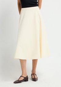 Vrouw in een crèmekleurige midi-rok met donkerbruine T-strap low heel schoenen, staand voor een effen witte achtergrond.