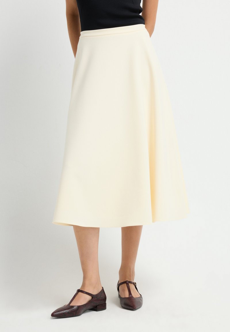 Vrouw in een crèmekleurige midi-rok met donkerbruine T-strap low heel schoenen, staand voor een effen witte achtergrond.
