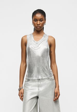 Kvinde med fletninger iført en ærmeløs sølvglitrende top og løse lysegrå bukser, stående mod en ensfarvet baggrund.