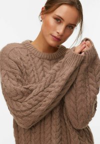 Femme aux cheveux châtain clair portant un pull taupe en maille torsadée épaisse avec de grandes manches, tenant ses mains près de son visage.