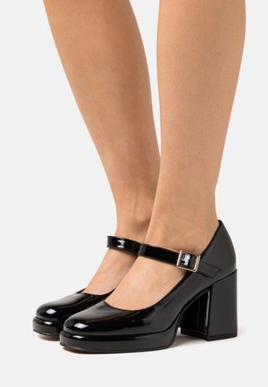 Beine in schwarzen Lackleder-Mary-Jane-Schuhen mit Blockabsätzen und Schnallenriemen vor einem schlichten weißen Hintergrund.