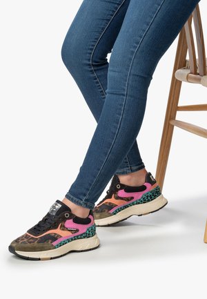 Persoon in blauwe skinny jeans en kleurrijke sneakers met luipaardprint, roze, oranje, teal en beige, zittend op een houten stoel.