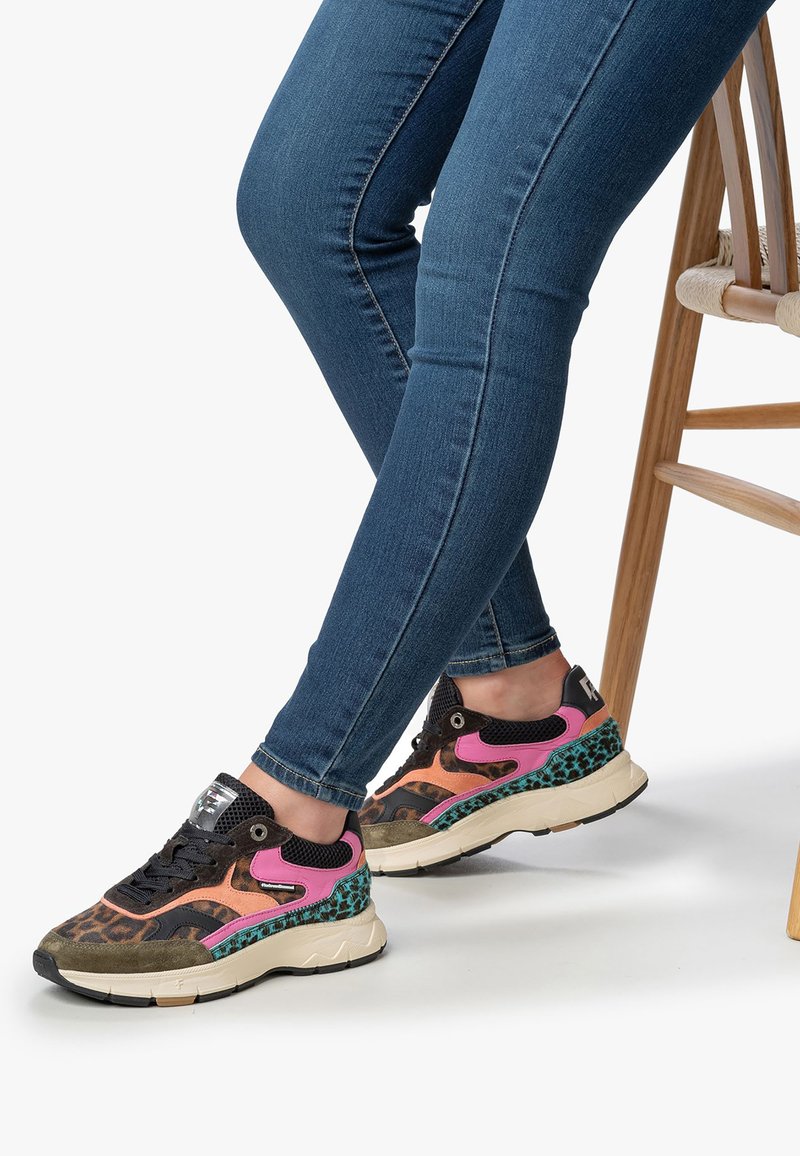 Persona che indossa jeans skinny blu e scarpe colorate con stampa leopardata, rosa, arancione, verde acqua e beige, seduto su una sedia di legno.