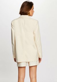 Blazer beige con pequeños lunares, con solapa de muesca, mangas largas y un corte relajado, combinado con pantalones cortos beige. Diseño minimalista.