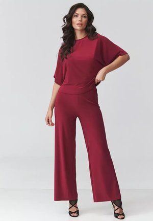 Kobieta z długimi ciemnymi włosami, ubrana w luźny bordowy top i szerokie spodnie, stoi z jedną ręką na biodrze, widoczne czarne sandały na obcasie z paskami.