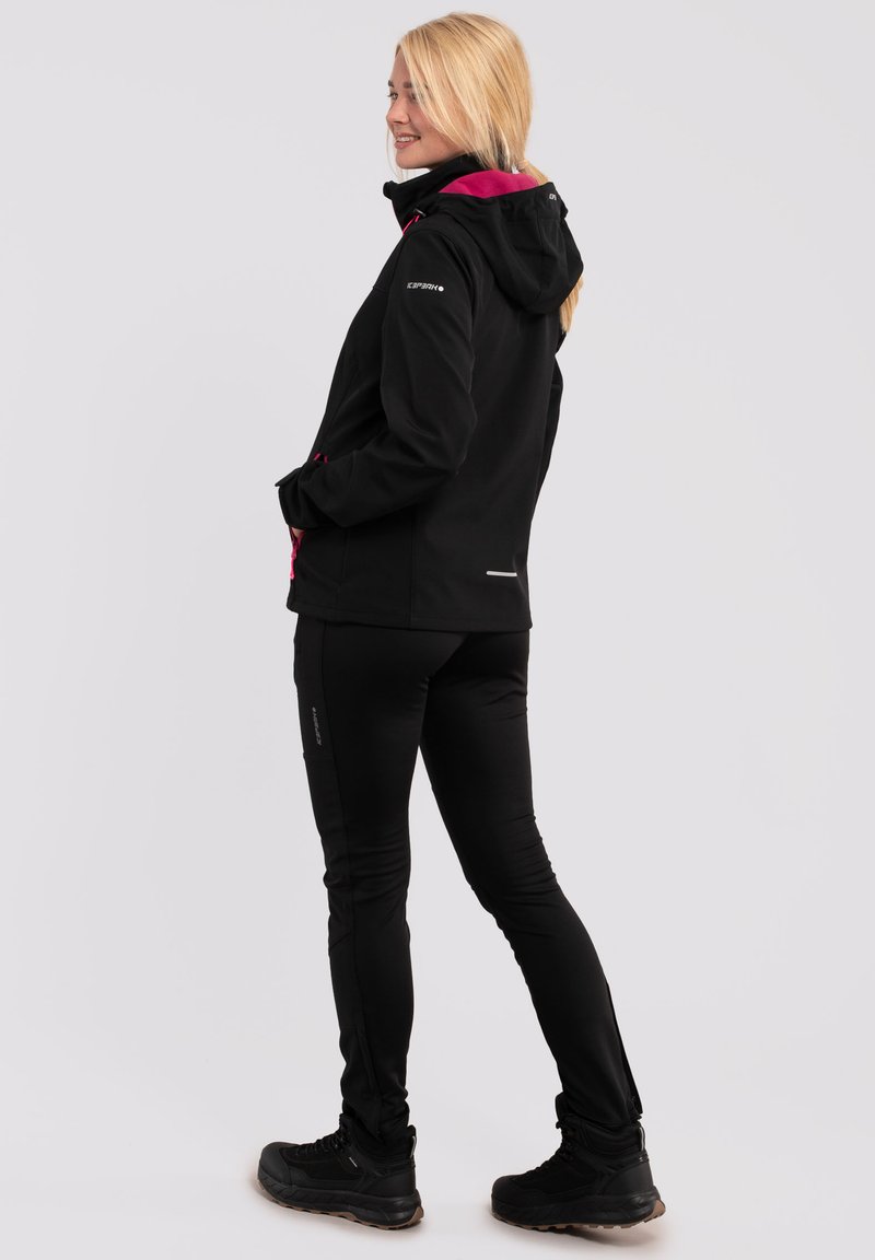 Giacca esterna nera con dettagli rosa, cappuccio e tasche con zip, abbinata a pantaloni neri aderenti e stivali da trekking neri.