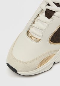 Beige lenkkari, jossa yhdistyvät sileä nahka ja mokkanahka, kultainen korostuslogo ja valkoiset kengännauhat, kaikki kiinnittynyt teksturoidulle mustalle pohjalle.