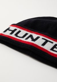 Fekete kötött sapka piros és fehér csíkkal, amelyen a "HUNTER" szó vastag betűkkel van feltüntetve. Puha tapintású, jól illeszkedő.
