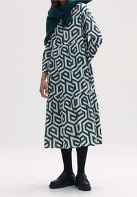 Långärmad klänning med ett geometriskt mönster i grönt och vitt, med en volangkjol och cinchad midja. Kombinerad med svarta loafers och strumpbyxor.