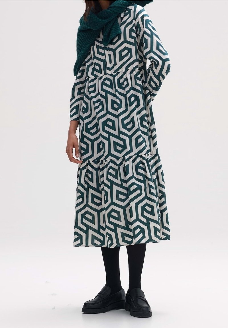 Långärmad klänning med ett geometriskt mönster i grönt och vitt, med en volangkjol och cinchad midja. Kombinerad med svarta loafers och strumpbyxor.