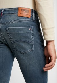 Närbild av en person som bär blå Tommy Jeans denimjeans med en brun läderlogg på bakfickan och en liten flagglogotyp.