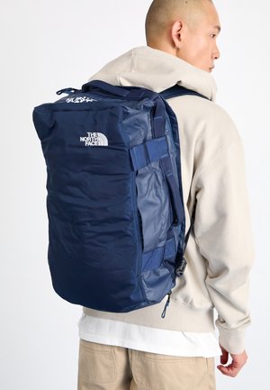 Ein Mann in einem beigen Hoodie und beiger Hose trägt einen großen dunkelblauen Rucksack von The North Face auf dem Rücken und steht seitlich vor einem weißen Hintergrund.