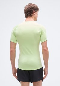 Αθλητική κοντομάνικη μπλούζα ανοιχτό πράσινο από ελαστικό ύφασμα, με εφαρμοστή γραμμή και λογότυπο στην πίσω περιοχή του λαιμού. Οι σορτς είναι μαύρα.