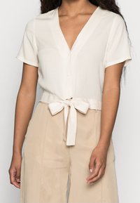 Mujer vestida con una blusa de cuarzo, de manga corta y escote en V, con botones y un lazo atado a la cintura, combinada con pantalones beige claros.