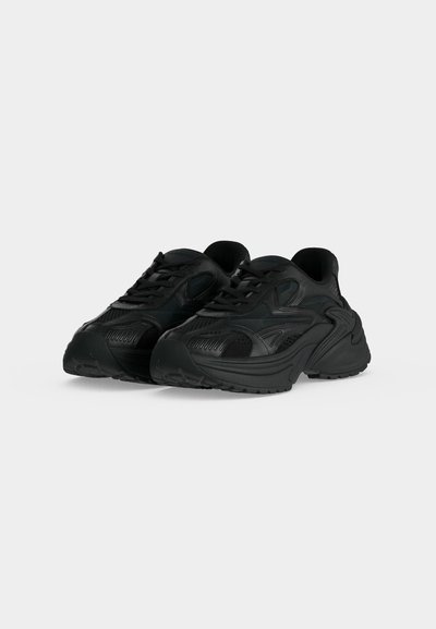 Ζεύγος μαύρα chunky sneakers με υφασμάτινες λεπτομέρειες και παχιές σόλες, εμφανίζεται σε απλό ανοιχτό γκρι φόντο.