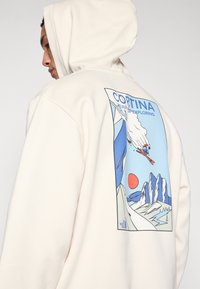 Krema kapuljača s grafičkim printom na leđima koji prikazuje snježne planine, skijaša i crveno sunce u plavim i bijelim tonovima.