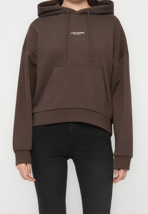Person iført en mørkebrun cropped hoodie med teksten "JJXX STUDIO" på brystet og sort tætsiddende bukser, stående mod en ensfarvet baggrund.