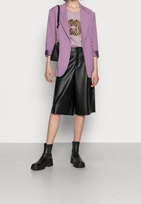 Blazer de lavanda, camiseta gráfica con labios de estampado animal, pantalones cortos de piel sintética negra y botas de tobillo negras de suela gruesa. Bolso negro simple.