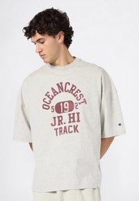 Jonge man met krullend haar kijkt naar beneden, gekleed in een lichtgrijze oversized T-shirt met "OCEANCREST JR. HI TRACK 192" in wijnrode tekst.