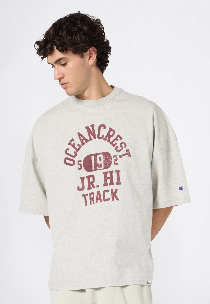 Jonge man met krullend haar kijkt naar beneden, gekleed in een lichtgrijze oversized T-shirt met "OCEANCREST JR. HI TRACK 192" in wijnrode tekst.