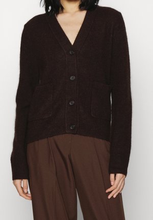 Personne portant un cardigan marron foncé boutonné avec des poches avant et un pantalon marron ample assorti sur un fond clair uni.
