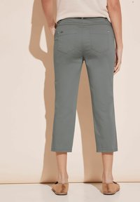 Kvinna iklädd grå cropped byxor och beiga platta skor, stående mot en beige vägg, sedd från ryggen.