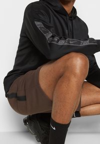 Svart huvtröja med grå Nike-logotyp på ärmarna, i kombination med bruna shorts med svarta sidostrikar, och svarta skor med synlig sula.