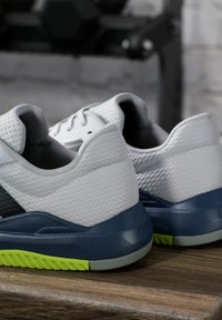 Zapatillas deportivas blancas con parte superior de malla texturizada, acentos en azul oscuro y una suela de goma verde y gris. Cuenta con un talón acolchado para mayor comodidad.